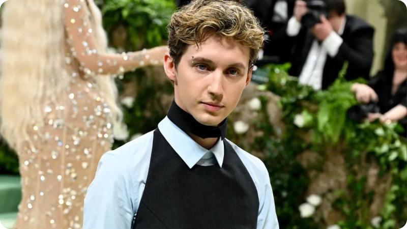 A young man with short, wavy hair is wearing a stylish outfit featuring a light blue shirt under a black vest with a unique black neck accessory. He is posing confidently at an event, with a blurred background of greenery and photographers capturing the moment. In the background, there is a glimpse of another person in a sparkling gown.