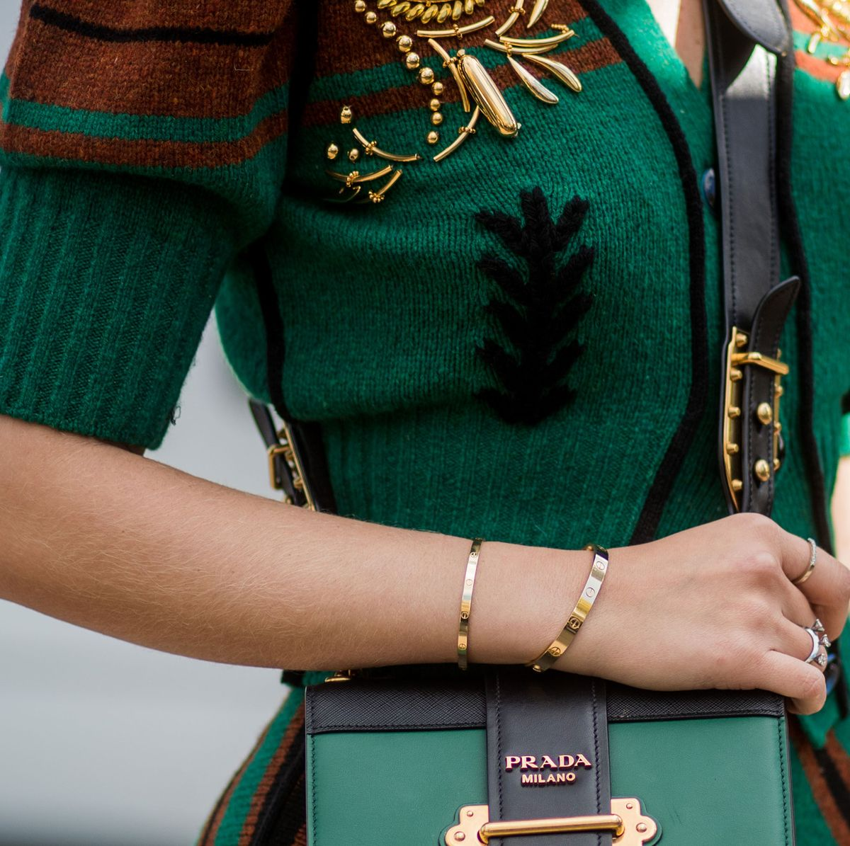 Close-up of a woman's outfit featuring a green and brown sweater with gold embellishments, a black and green Prada Milano handbag, and a gold bracelet on her wrist.