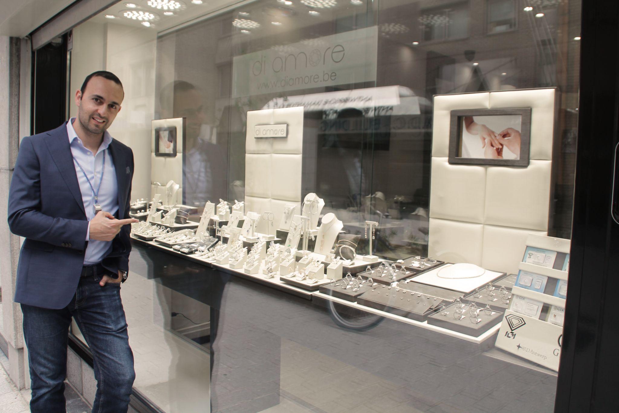 Owner of Di Amore jewelry store pointing to the window display showcasing a variety of elegant jewelry pieces, with the Di Amore logo and website visible in the reflection.