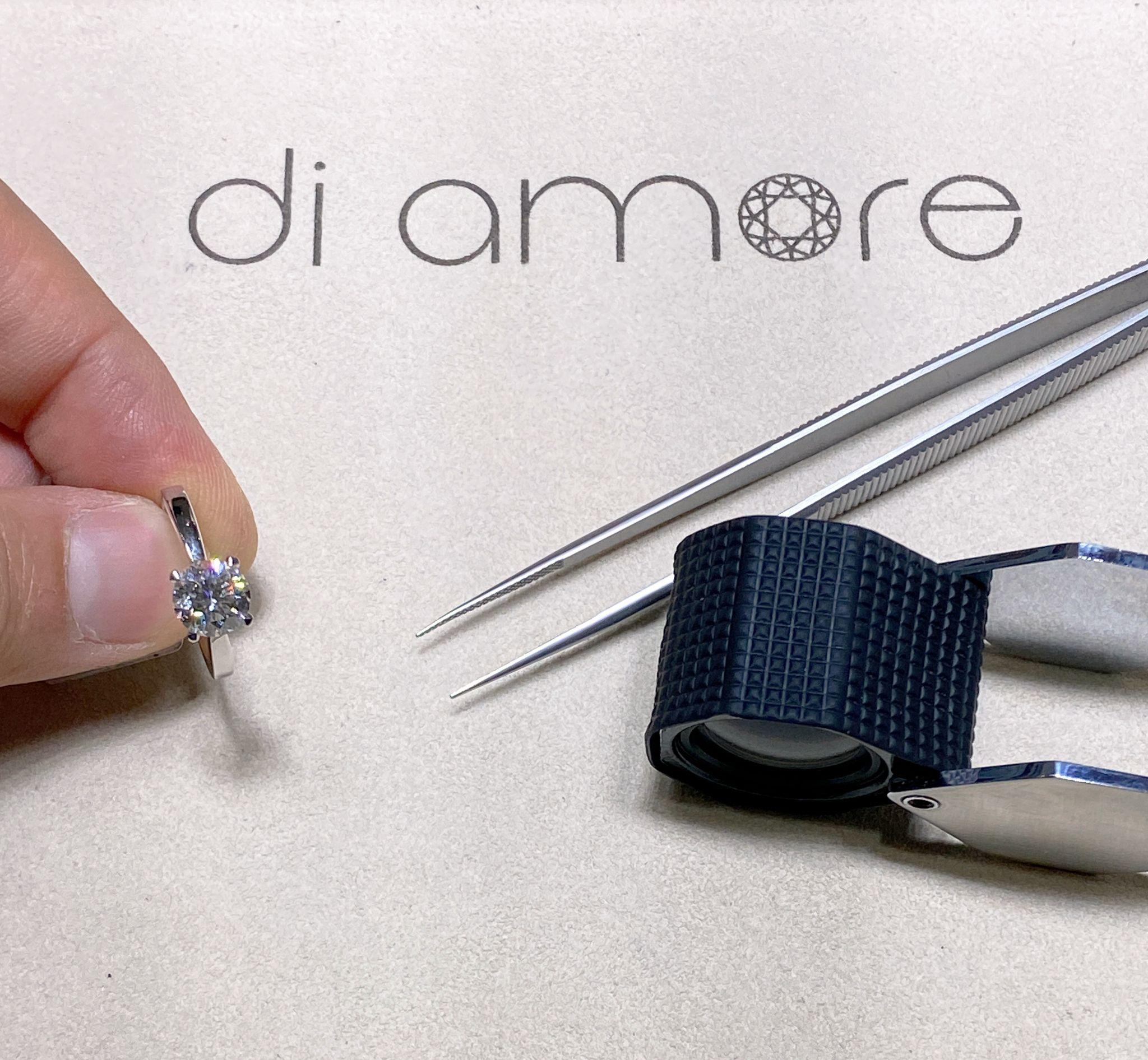 Hand holding a diamond ring next to jeweler's tools including tweezers and a loupe, with the Di Amore logo visible in the background.
