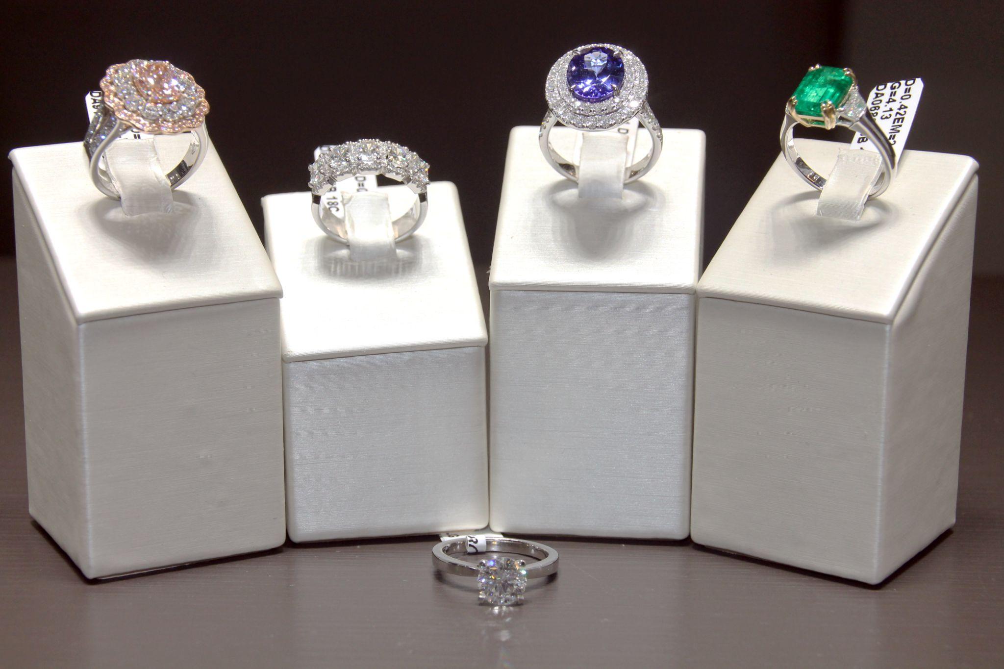 Vijf elegante ringen met verschillende edelstenen, waaronder een roze diamant, witte diamanten, een blauwe saffier en een groene smaragd, gepresenteerd op zilverkleurige standaards.
