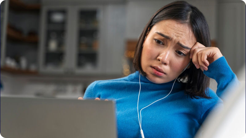 Woman in a blue sweater looking worried while working on a laptop, with earphones in, sitting in a home environment.
