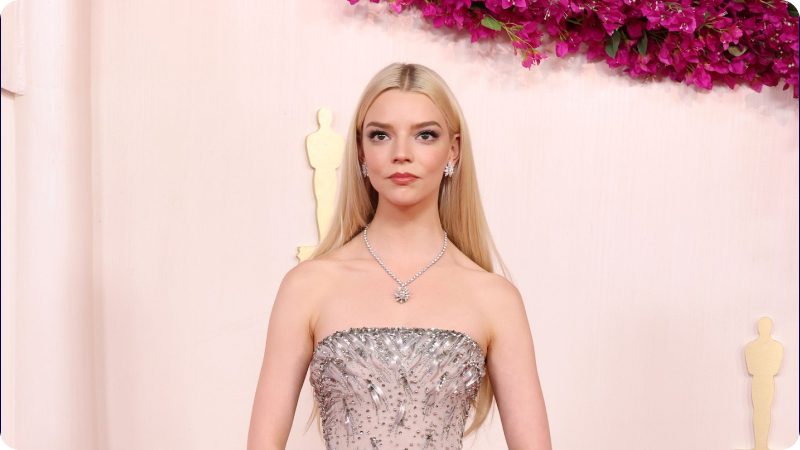 Anya Taylor-Joy wearing a shimmering strapless gown and elegant jewelry, posing at an event or awards ceremony with Oscar statuettes and pink flowers in the background.