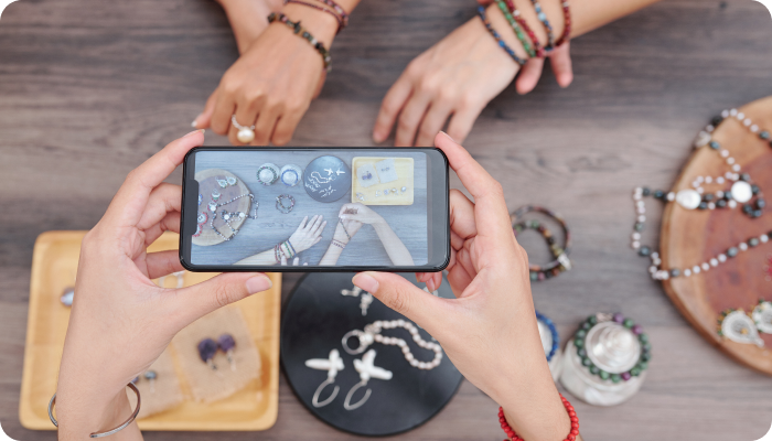 Hands holding a smartphone taking a photo of various pieces of jewelry displayed on a table, with additional hands arranging the jewelry, capturing the process of showcasing and organizing jewelry for presentation.