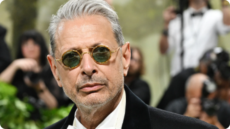 A man with silver hair is wearing round, tinted sunglasses and a black velvet suit with a white shirt. He has a confident, slightly serious expression. The background shows a blurred crowd of photographers and attendees, highlighting the formal and glamorous atmosphere of the event.