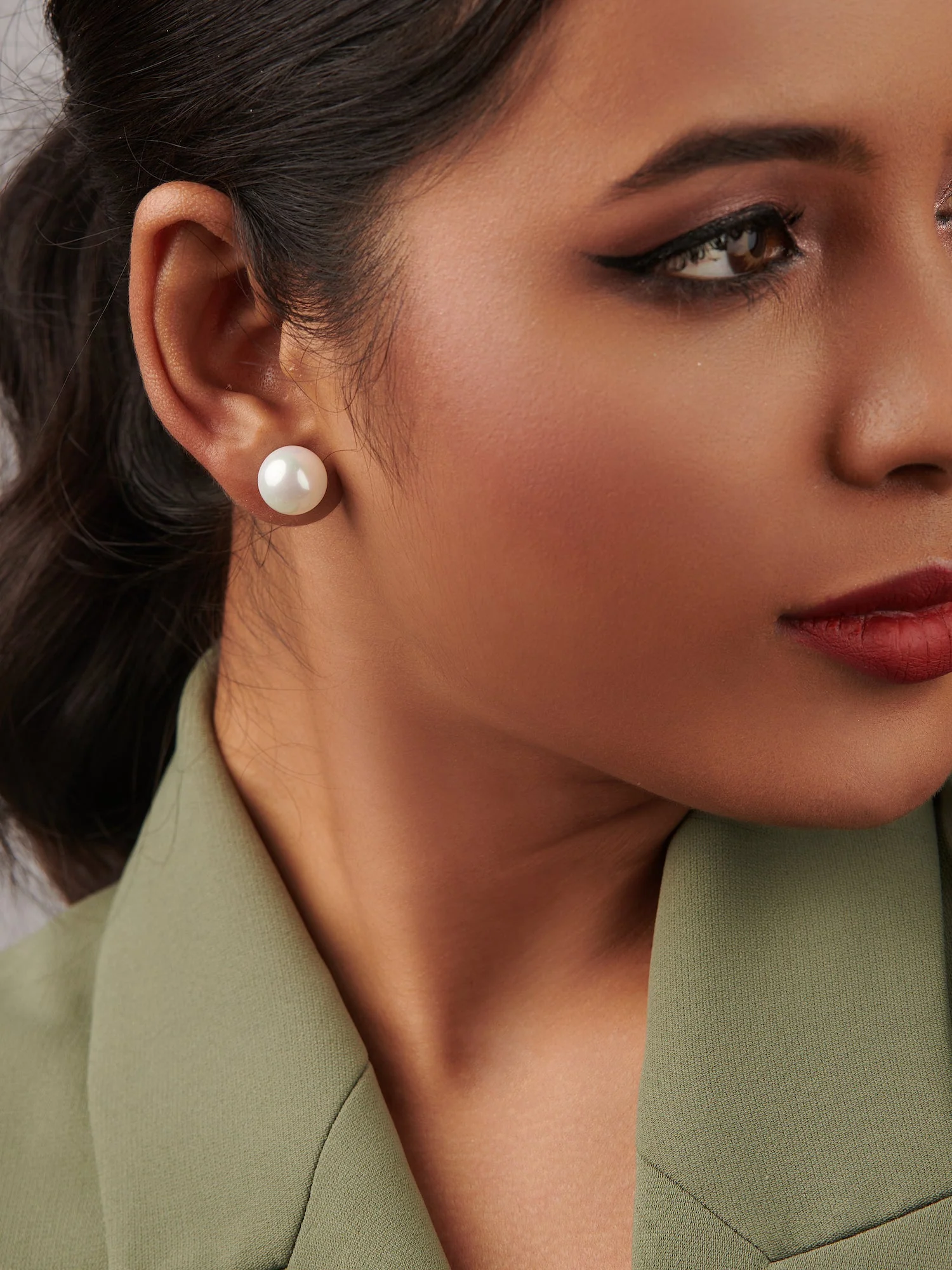 Close-up of a woman wearing pearl stud earrings, with her hair styled in a low ponytail, and wearing a green blazer.