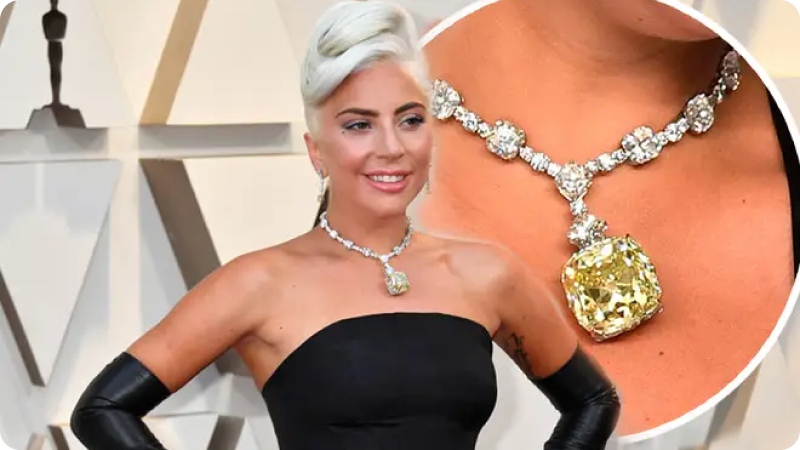 "Lady Gaga wearing a black strapless gown and long black gloves, showcasing an extravagant diamond necklace with a large yellow diamond pendant at an awards ceremony.