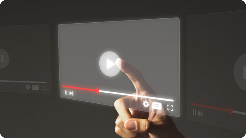 A hand interacting with a virtual video player interface, pressing the play button. The interface includes typical video player controls such as play/pause, a progress bar with a red indicator, volume control, settings, and fullscreen options. The background is dark, highlighting the translucent, glowing elements of the video player interface.