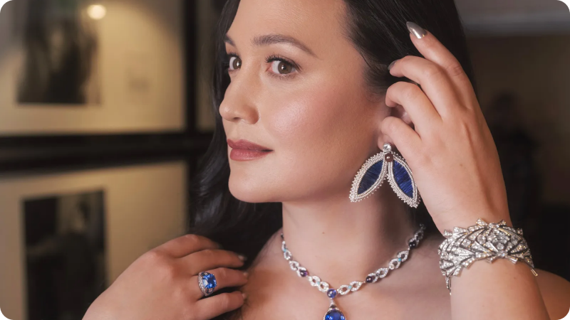 A woman showcasing luxurious jewelry, including large blue gemstone earrings, a matching necklace, a ring, and a diamond bracelet, highlighting elegance and sophistication.