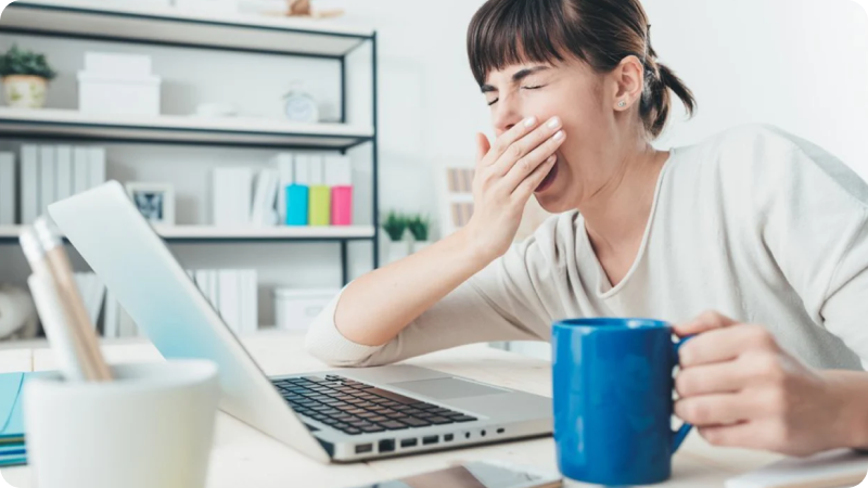 Woman sitting at a desk with a laptop, yawning and holding a blue coffee mug.