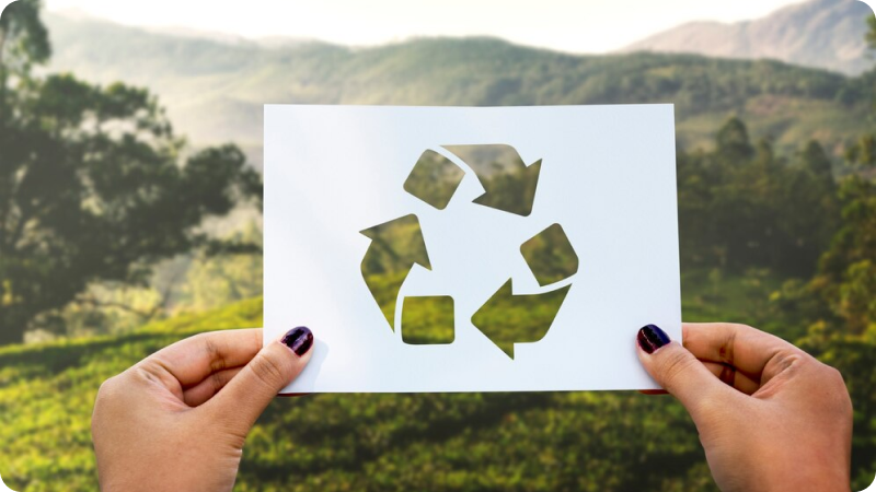 Hands holding a paper with a cut-out recycling symbol against a backdrop of a lush, green landscape.