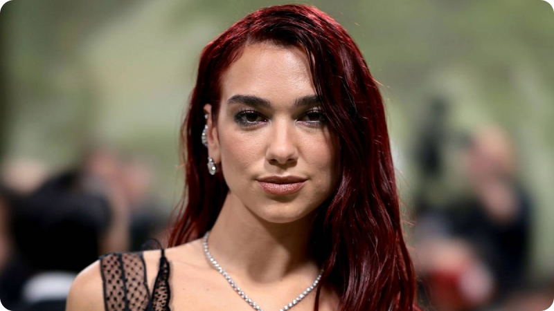A woman with long, dark red hair is wearing a subtle, elegant necklace and earrings. She has a confident expression and is dressed in a stylish outfit with delicate, lacy details. The background is blurred, highlighting the focus on her and the sophisticated ambiance of the event.