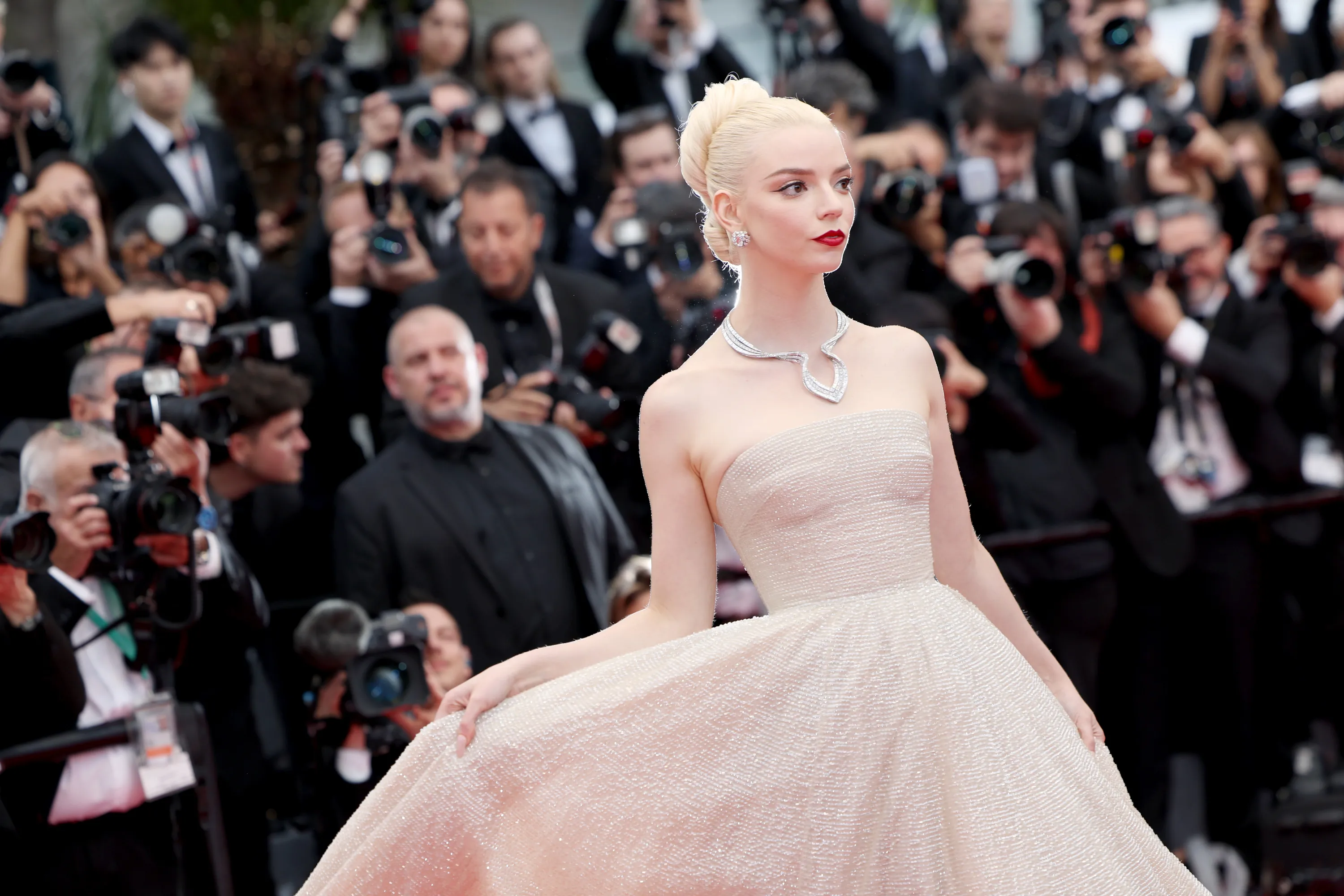 Woman with platinum blonde hair in an elegant updo, wearing a strapless shimmering gown and statement necklace, walking the red carpet with photographers in the background capturing the moment.