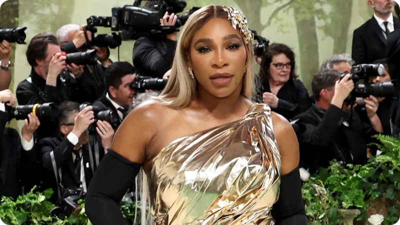 A woman with long, blonde hair is wearing a metallic gold, one-shoulder gown paired with long black gloves. She is accessorized with a jeweled hairpiece and matching earrings. The background features a crowd of photographers and event attendees, emphasizing the glamorous and high-profile nature of the event.