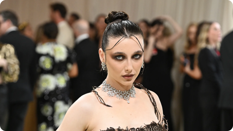 A woman with dark, sleek hair styled in an updo and accented with delicate strands framing her face is wearing a dark, intricate gown. She has dramatic, smoky eye makeup and is adorned with a detailed choker necklace and earrings. The background shows a blurred crowd of event attendees, adding to the sophisticated and glamorous atmosphere
