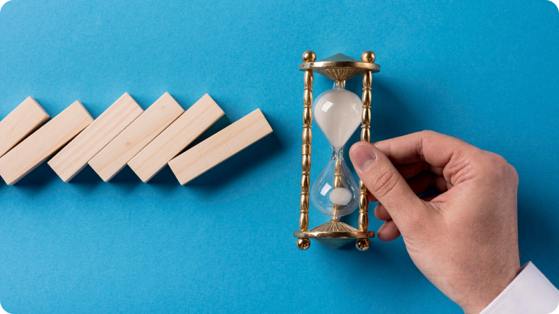 Hand holding an hourglass next to a row of wooden dominoes against a blue background