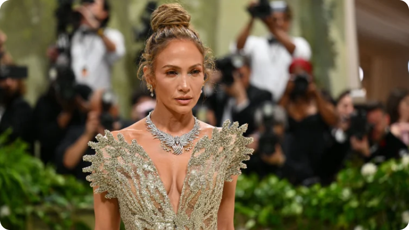 A woman dressed in an elegant, shimmering gown with intricate leaf-like details, wearing a striking necklace and earrings, with her hair styled in an updo. She is posing on a green, garden-themed carpet, with photographers and onlookers in the background capturing the moment