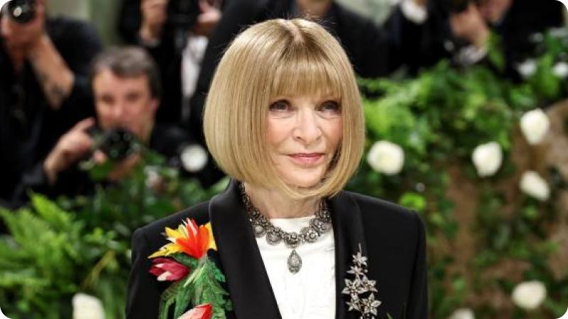 A woman with a short, blonde bob hairstyle is wearing a black blazer adorned with a colorful floral decoration and a white blouse. She is accessorized with a statement necklace and a jeweled brooch. The background features a lush, green setting with photographers capturing the event, highlighting the sophisticated and high-profile atmosphere.