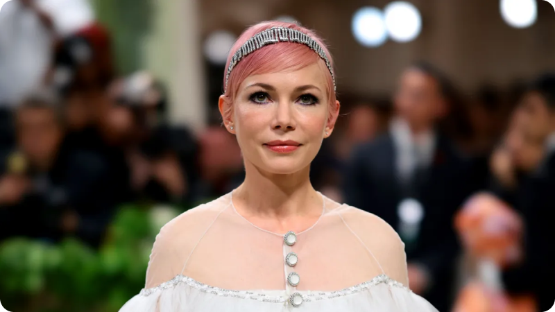 A woman with short, pink hair is wearing an elegant white dress with sheer details and decorative buttons down the front. She is accessorized with a sparkling headband and delicate earrings. The background features a blurred crowd of event attendees and photographers, highlighting the sophisticated and glamorous atmosphere of the event.