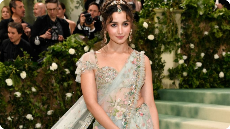 A woman with dark, wavy hair styled in an intricate updo is dressed in a stunning, embroidered gown with floral details and a sheer, flowing cape. She is wearing matching jewelry, including earrings and a headpiece. The background features a lush, green setting with photographers and onlookers, highlighting the elegance and sophistication of the event.