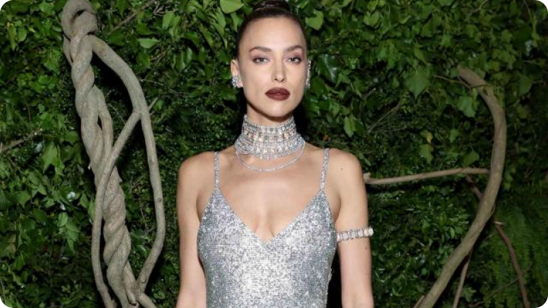 A woman with her hair styled in a sleek updo is wearing a sparkling silver gown with thin straps and intricate embellishments. She is adorned with a dramatic choker necklace, matching arm cuff, and earrings, and has bold, dark lipstick. The background features lush green foliage, creating a striking contrast with her glamorous outfit.