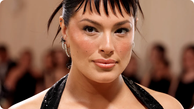 A close-up of a woman with dark hair styled in a sleek updo with short, wispy bangs. She is wearing a shimmering black halter dress and sparkling earrings. Her makeup is polished, highlighting her natural beauty. The background is softly blurred, emphasizing her radiant and confident expression.