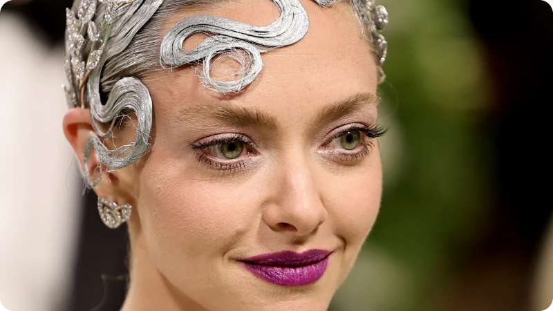 A close-up of a woman with intricate silver designs in her hair, wearing dramatic purple lipstick and delicate, sparkling earrings. She has a serene expression and a radiant complexion. The background is softly blurred, focusing on her detailed and artistic hairstyle and makeup, highlighting the elegant and creative atmosphere of the event.