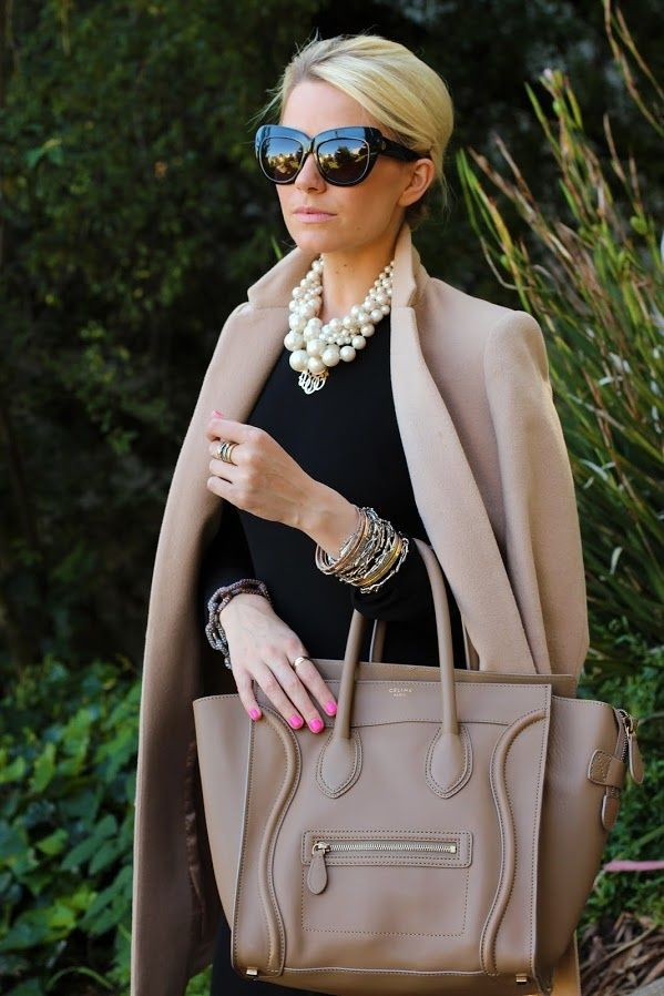 Fashionable woman wearing sunglasses, a beige coat, and a black dress with a large pearl necklace and multiple bracelets, holding a beige designer handbag, posing outdoors
