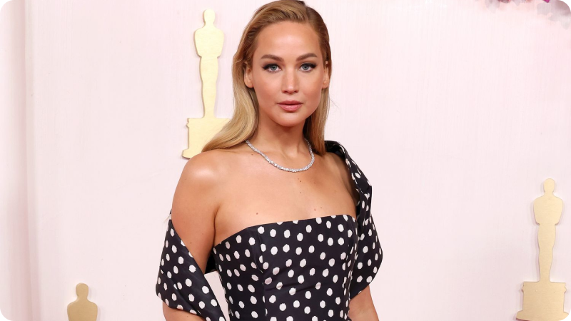 Jennifer Lawrence wearing a stylish black and white polka dot gown and a diamond necklace, posing at an awards ceremony with Oscar statuettes in the background