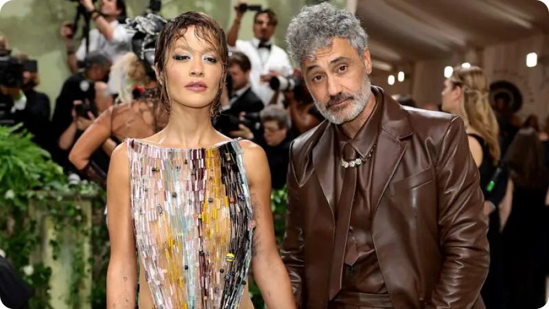 A woman and a man are posing together at a high-profile event. The woman is wearing a sleeveless, multicolored, mosaic-patterned gown with wet-look hair and dramatic makeup. The man is dressed in a brown leather suit with a matching shirt and tie, accessorized with a necklace. The background features other attendees and photographers, emphasizing the glamorous and fashionable atmosphere of the event.