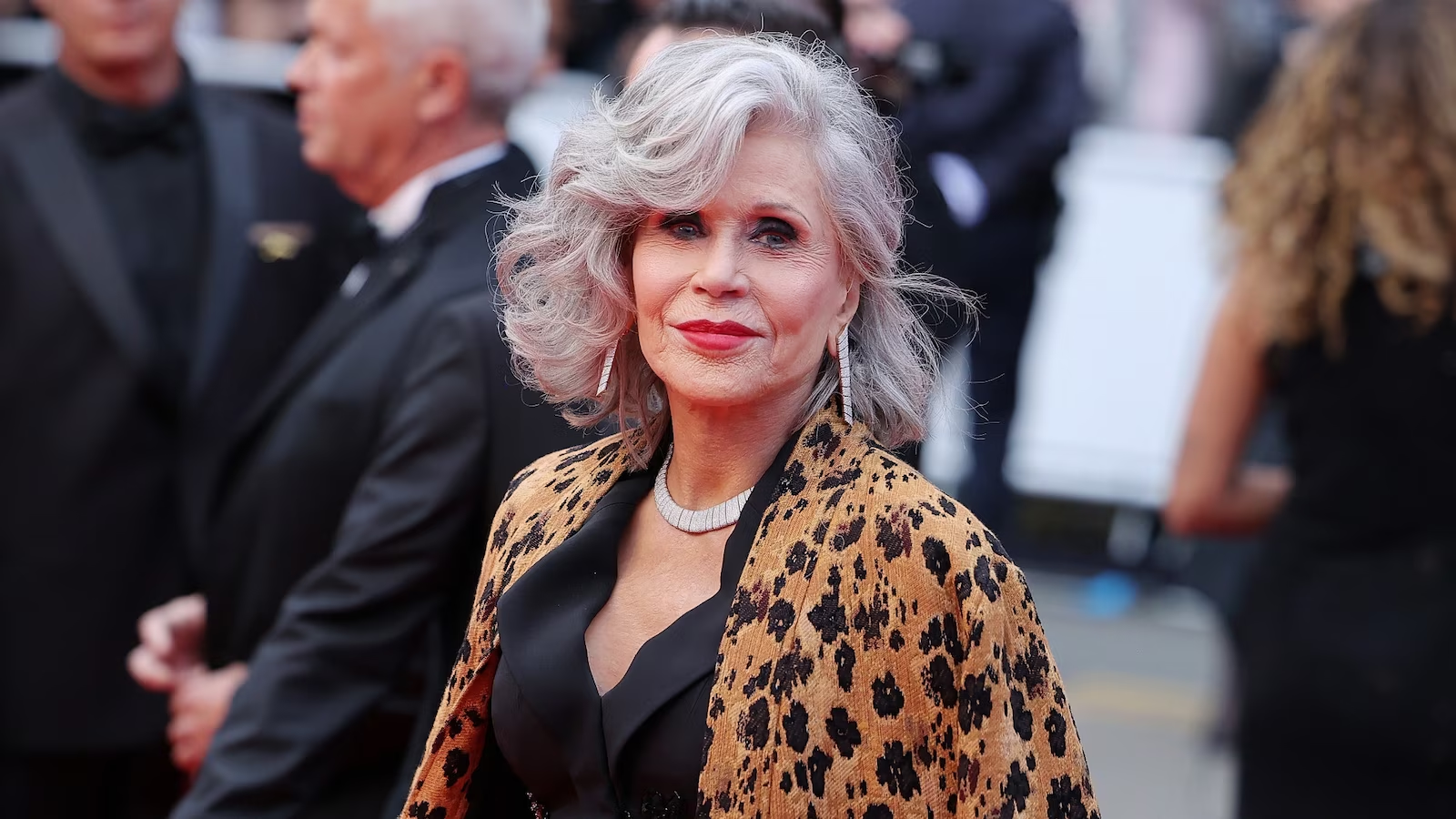 Jane Fonda with styled silver hair in a black outfit and leopard-print coat, accessorized with statement earrings and necklace, on a red carpet at a formal event.