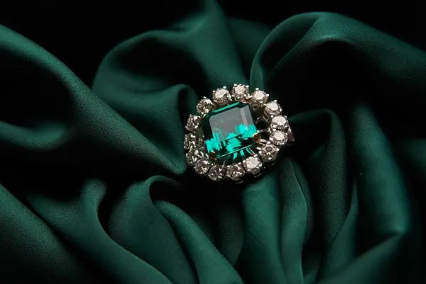 Close-up of a luxurious ring with a large emerald-cut green gemstone surrounded by a halo of sparkling diamonds, placed on a rich dark green fabric.