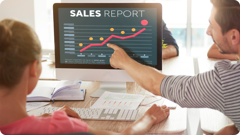 Two people reviewing a sales report displayed on a computer monitor, showing a rising graph and data points, indicating an increase in sales performance