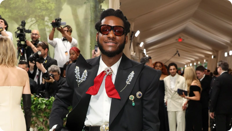 A man wearing a stylish black suit adorned with decorative brooches and floral details on the lapels. He is accessorized with a red bow tie, a white shirt, and round sunglasses. The background features other event attendees and photographers, emphasizing the fashionable and sophisticated atmosphere of the event.