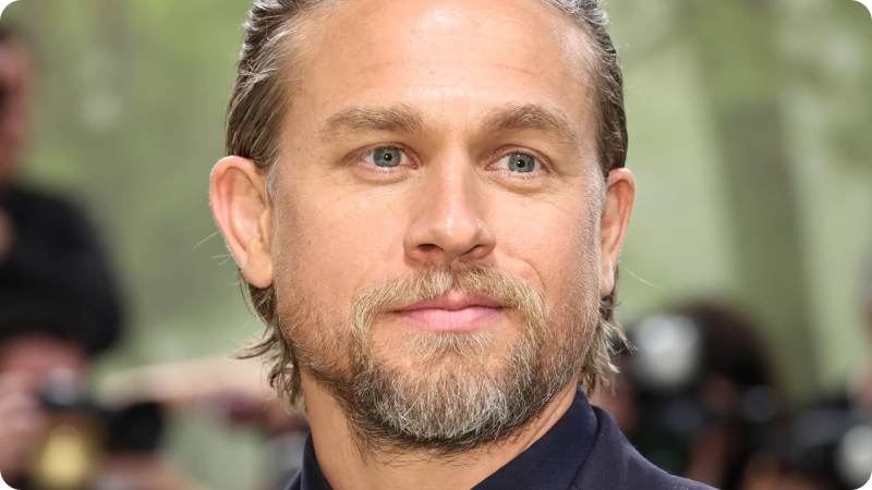 A close-up of a man with neatly groomed facial hair and slicked-back hair. He is wearing a dark suit and has a calm, confident expression. The background features a blurred crowd of photographers and event attendees, highlighting the sophisticated and elegant atmosphere of the event.