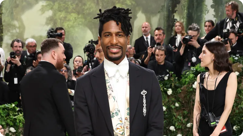 A man dressed in an elaborate, vintage-inspired outfit featuring a velvet coat, a white cravat, and a top hat. He is posing with one hand holding the brim of his hat. The background consists of lush green foliage, creating a dramatic and elegant atmosphere. give me alt tags