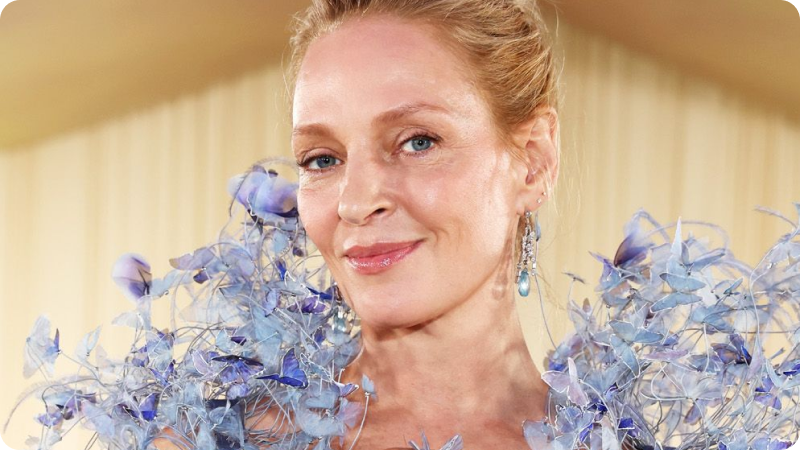A woman with her hair pulled back is wearing a dress adorned with delicate, blue, floral-like details that surround her shoulders. She has a serene, confident expression and is wearing elegant drop earrings. The background features a soft, beige setting, highlighting her radiant and sophisticated appearance.