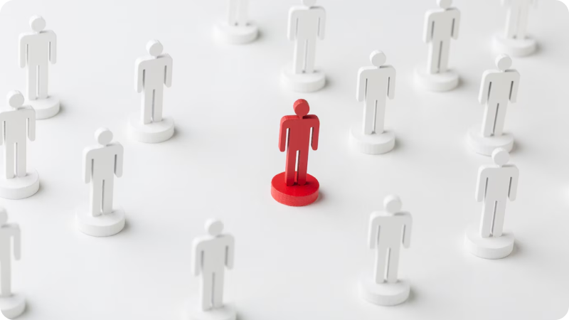 Several white human figures standing on a white surface, with one red figure in the center, symbolizing individuality or uniqueness. The arrangement emphasizes the contrast between the red figure and the white figures, highlighting the concept of standing out from the crowd or being different. The background is plain and white, focusing attention on the figures.