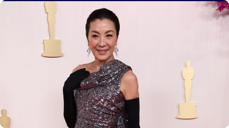 Michelle Yeoh wearing a glamorous, sparkling gown and black gloves, smiling confidently at an awards ceremony with Oscar statuettes in the background
