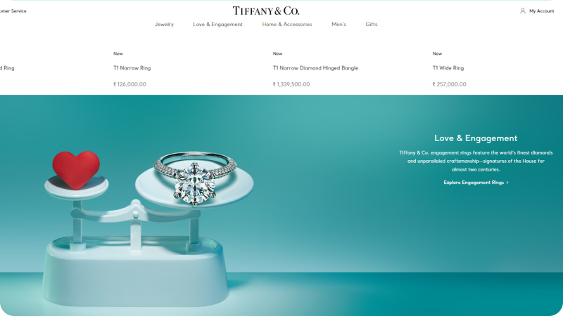A Tiffany & Co. webpage showcasing a balance scale with a red heart on one side and a diamond engagement ring on the other, highlighting the 'Love & Engagement' collection with prices listed for various jewelry items.
