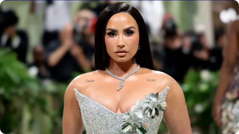 A woman with long, dark hair is wearing a shimmering, form-fitting silver gown with a deep neckline and floral embellishments. She is accessorized with a diamond necklace and has a confident expression. The background features a blurred crowd of photographers and attendees, emphasizing the glamorous atmosphere of the event.
