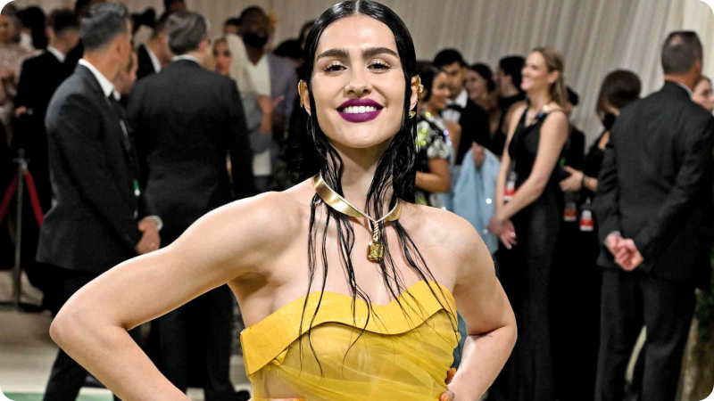 A woman with long, dark, wet-look hair is wearing a vibrant yellow strapless gown. She has a bold, dark purple lipstick and is accessorized with a unique gold necklace. She is smiling confidently with her hands on her hips. The background shows a crowd of other attendees, emphasizing the lively and glamorous atmosphere of the event.