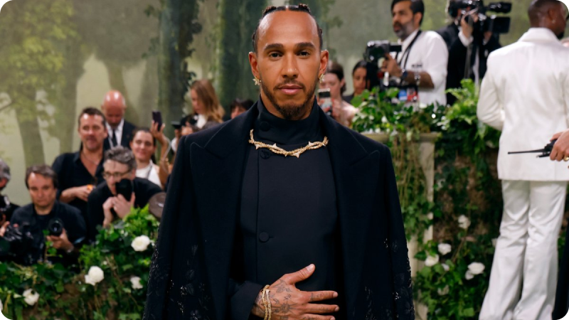 A man with a close-cropped hairstyle is dressed in an elegant, all-black outfit featuring a high-neck top and a long coat with intricate details. He is wearing a distinctive gold necklace and has a calm, composed expression. The background features a lush, green setting with photographers and attendees, adding to the sophisticated ambiance of the event.