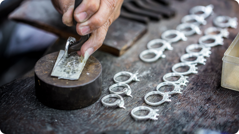 A craftsman working on a piece of jewelry at a workbench, with several unfinished ring mounts arranged neatly beside the tools.