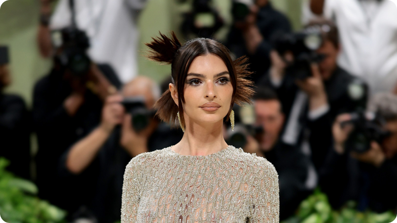A woman with dark hair styled in an updo with spiky, feather-like details is wearing an intricately beaded, sheer gown. She has a poised expression and is accessorized with elegant drop earrings. The background features photographers and other event attendees, emphasizing the sophisticated and glamorous atmosphere of the event.