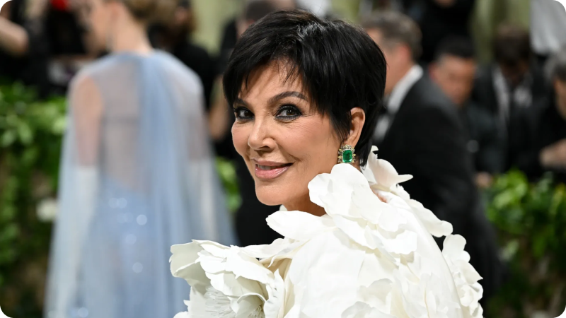 A woman with short, dark hair is smiling while wearing a white outfit adorned with large, ruffled floral details. She is accessorized with striking green earrings. The background features other event attendees and photographers, highlighting the elegant and glamorous atmosphere of the event.