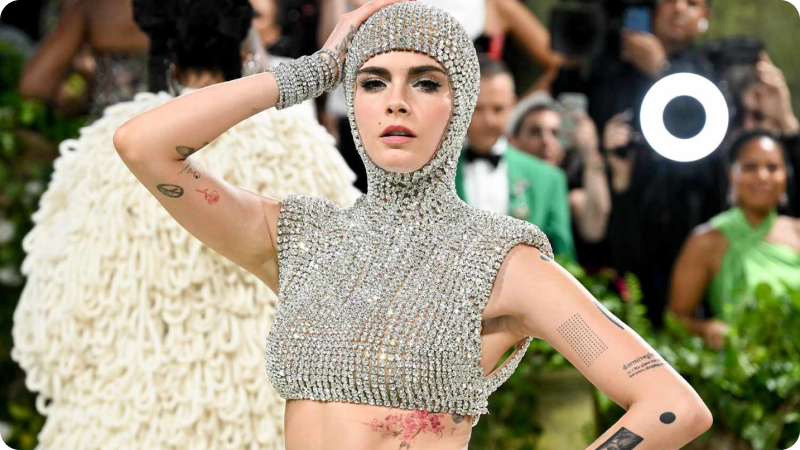 A woman with short, dark hair is wearing a sparkling, jeweled headpiece and matching crop top, revealing her midriff. She has various tattoos on her arms and torso. The background features other event attendees and photographers, highlighting the glamorous and fashionable atmosphere of the event.