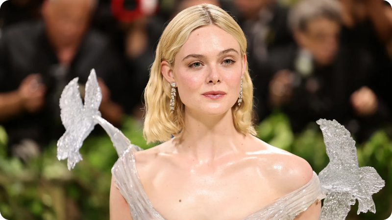 A woman with shoulder-length blonde hair is dressed in a delicate, shimmering gown with translucent, wing-like details on her shoulders. She is wearing elegant drop earrings and has a natural, radiant makeup look. The background shows a blurred crowd of photographers and onlookers, highlighting the event's high-profile nature