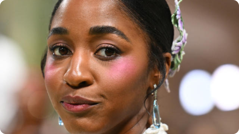 A close-up of a woman with dark hair styled back, adorned with a floral hair accessory. She has a natural, radiant complexion with a hint of pink blush on her cheeks and is wearing delicate drop earrings. The background is blurred, focusing on her expressive eyes and serene expression, highlighting the sophisticated atmosphere of the event.