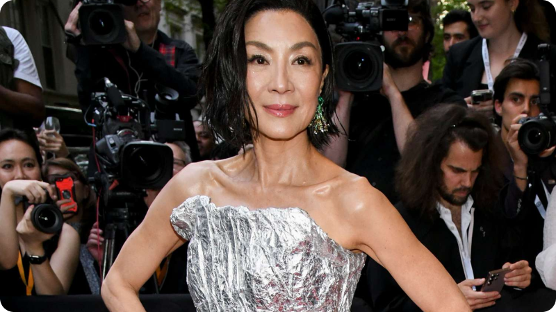 A woman with short, styled dark hair is wearing a striking, metallic silver gown with a unique, textured design. She is adorned with green statement earrings and has a confident expression. The background features a crowd of photographers and onlookers, emphasizing the event's high-profile nature and glamorous atmosphere